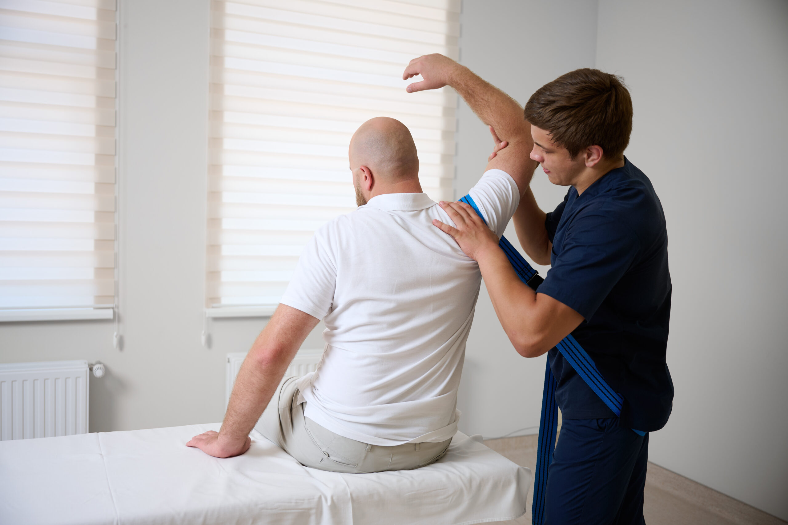 Chiropractor adjusting male patient's shoulder in clinic. Physiotherapist performing manual therapy to relieve pain, improve mobility, support rehabilitation after injury or musculoskeletal disorder.